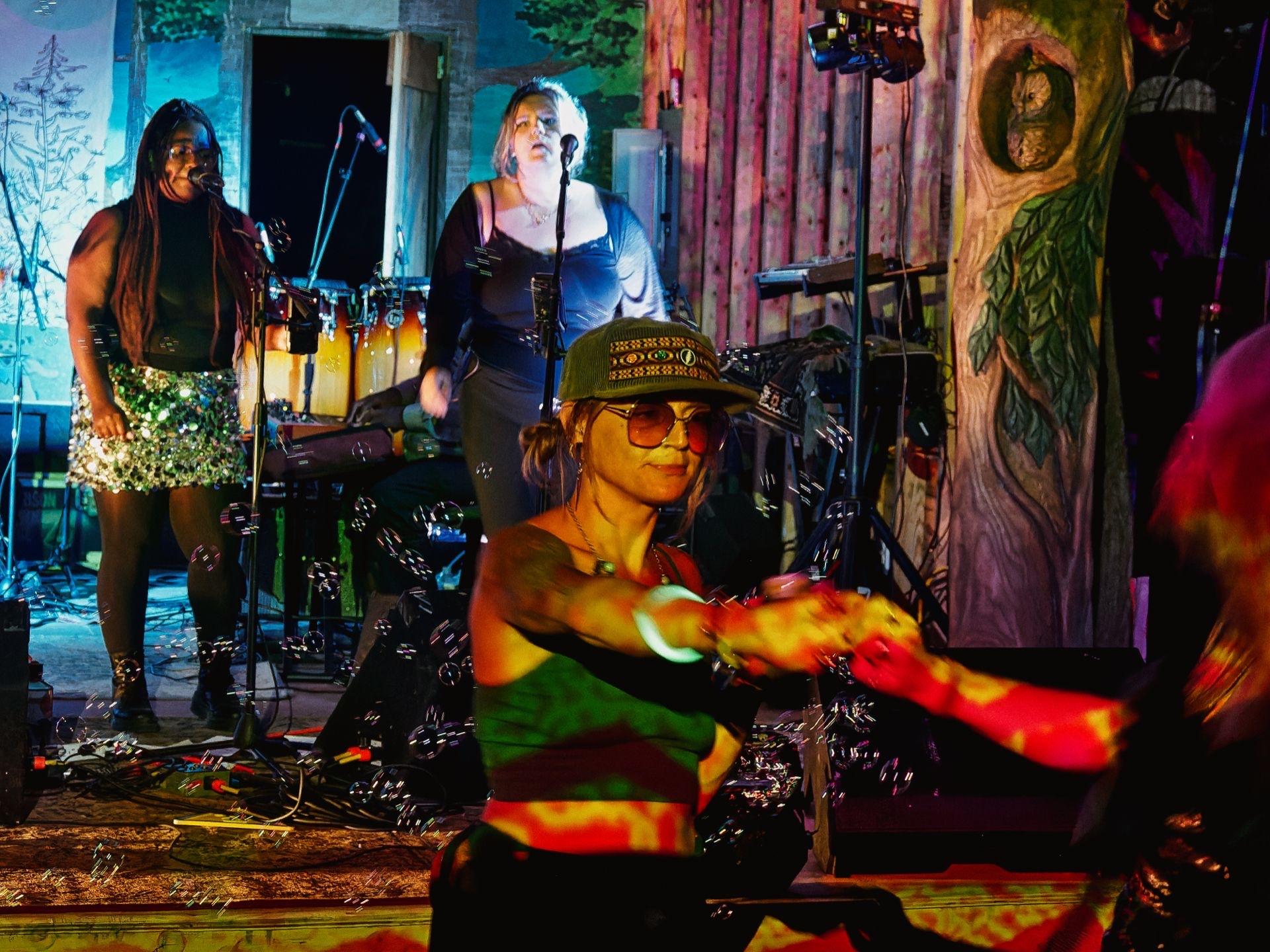 Live music performance in a warmly lit venue with painted woodland murals — a tree with an owl carved into it visible on the right. In the foreground, a performer in a green patterned cap and tinted sunglasses plays a tambourine with motion-blurred arms, bathed in red and green light. Behind her to the left, a woman in a sequined skirt sings into a microphone, and further back, a second singer in a blue top performs at her own mic beside a set of congas.