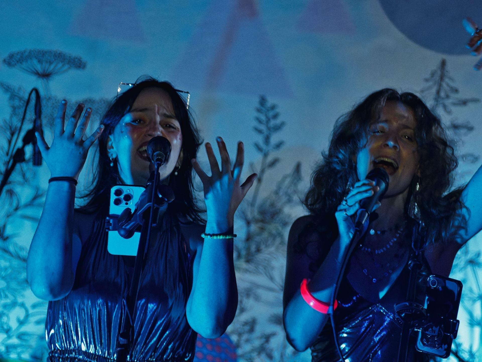 Two performers singing into microphones under deep blue stage lighting, both with hands raised and mouths open mid-song — the one on the left in a silver metallic top with a phone clipped to her micstand, the one on the right wearing a glowing red wristband. A botanical illustrated backdrop of silhouetted plants and trees fills the background.