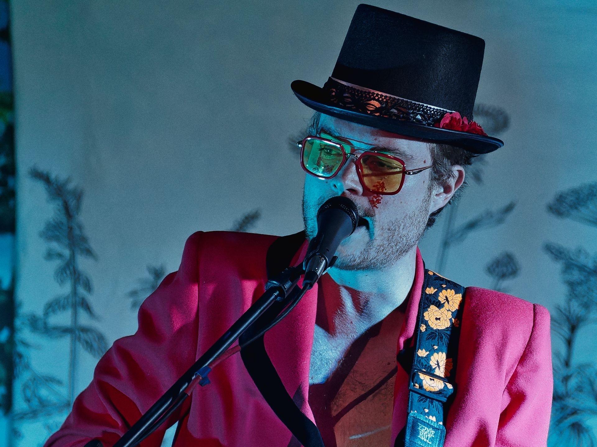 A performer in a red blazer, black top hat with lace trim, and oversized tinted aviator sunglasses sings into a microphone, a floral guitar strap crossing his chest. The background is a pale blue-washed curtain with shadowed tropical leaf prints, lit with cool stage lighting that gives the scene a moody, theatrical feel.