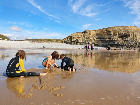 Picture of three children in wetsuits and swimming costumes playing in the sand. Background is a sandy beach, with a shingle slope and cliffs.