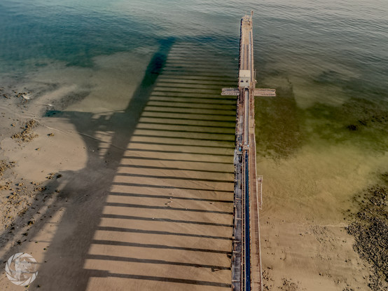 Photograph from a drone of a jetty extending out over sand and shallow sea water at low tide. Taken not long after sunrise, the shadow of the jetty extends to the left and is well defined on the wet sand and shallow water