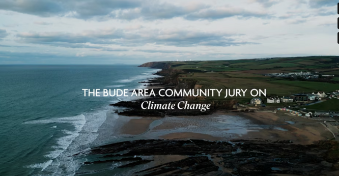 An aerial landscape photo of Bude, Cornwall, with the sea on the LHS, beach and coastline in the middle, and town and hills on the RHS.