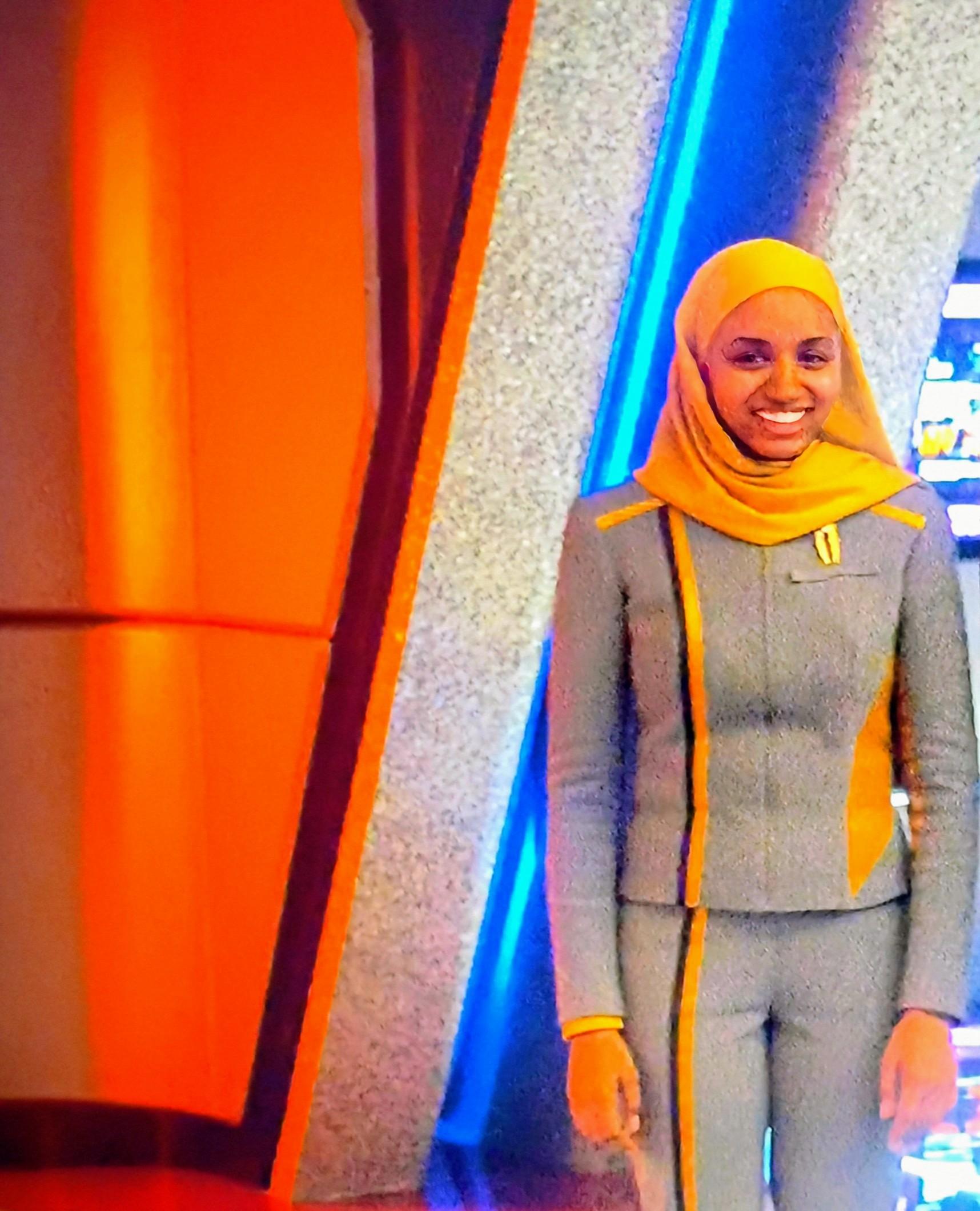 A Starfleet Academy engineering cadet is looking forward and smiling. The woman is wearing a yellow hijab to match her yellow-accented uniform. Starfleet Academy Episode 8.