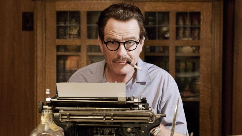 Bryan Cranston is sitting behind a typewriter. He has a pipe hanging from his mouth and has a quizzical look on his face.