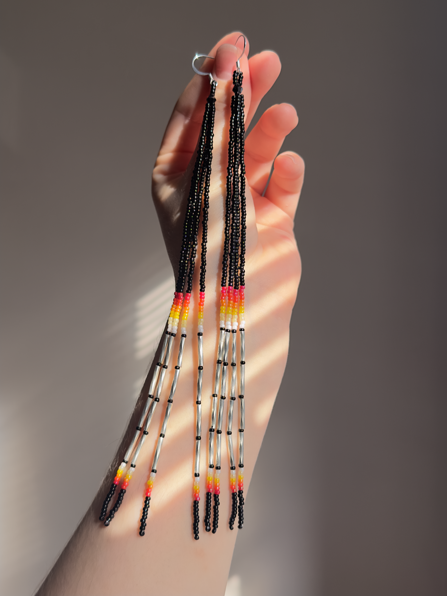 My hand holding a pair of long black beaded dangle earrings with red, orange, and yellow accents and silver bugle beads