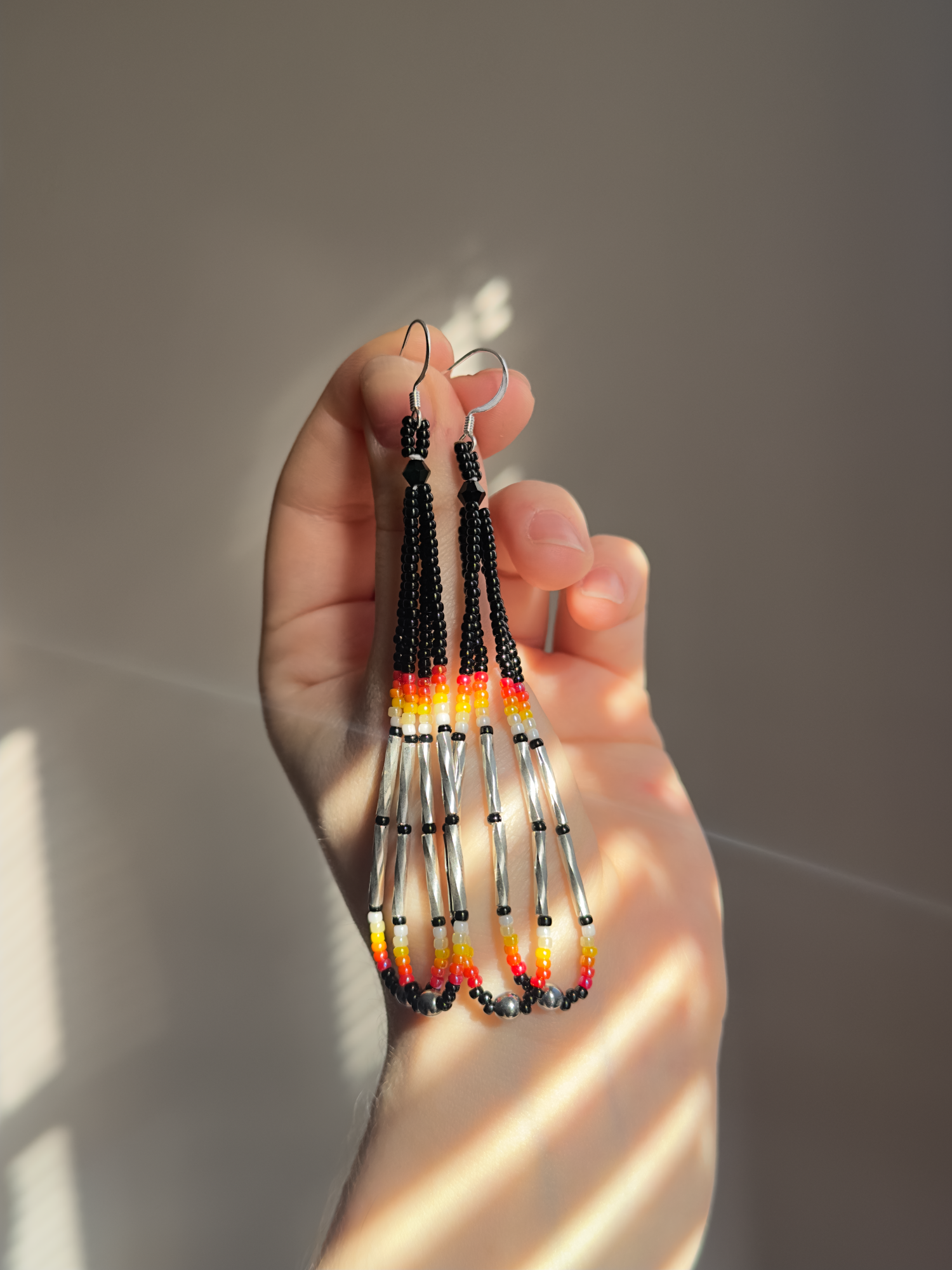 My hand holding a pair of black beaded loop earrings with red, orange, and yellow accents and silver bugle beads