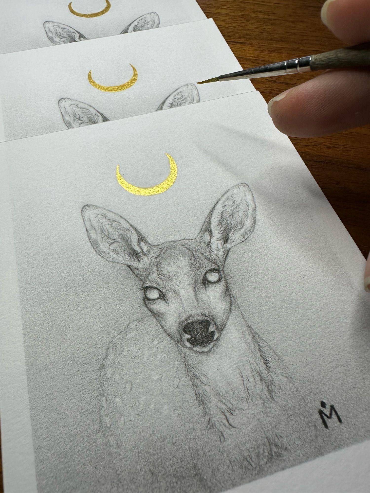 Studio shot of a print of my pencil drawing Moon Fawn. A young deer emerging from the mist. A gold crescent moon above her head.