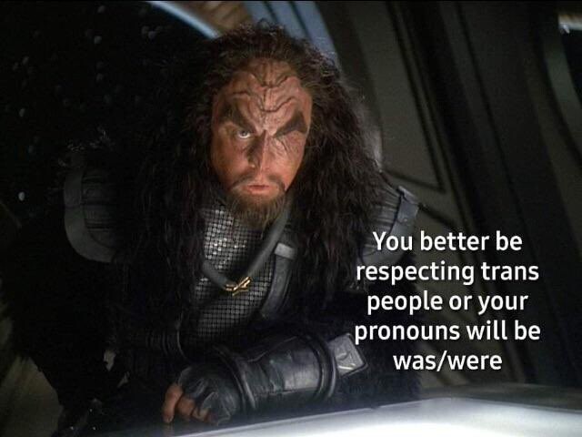 A Klingon warrior leaning over a bar, one of his eyes is missing and he has a stern expression on his face as he says "You better be respecting trans people or your pronouns will be was/were"