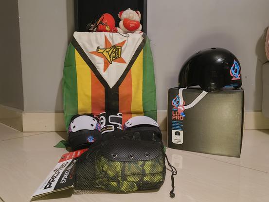 All the skate safety equipment laid out on the floor with a Zimbabwe flag in the background being helped up by cute teddies. There is one set of kneepads with the trans colours, and a second set with green camo. The helmet is placed on top of its box.