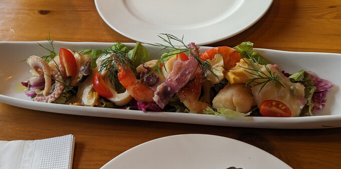 a narrow white dish with several types of seafood arranged on it. squid, shrimp, scallops, and a few others I can't identify. enough to be an appetizer for two people.
