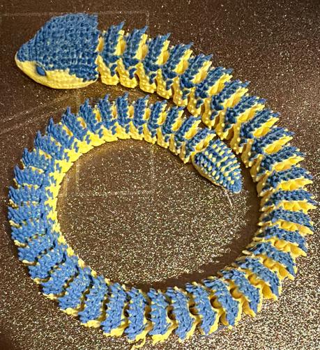 A 3D printed articulated snake in blue and yellow with blue eyes. The snake is resting on the gold colored, textured plate of the printer. 
