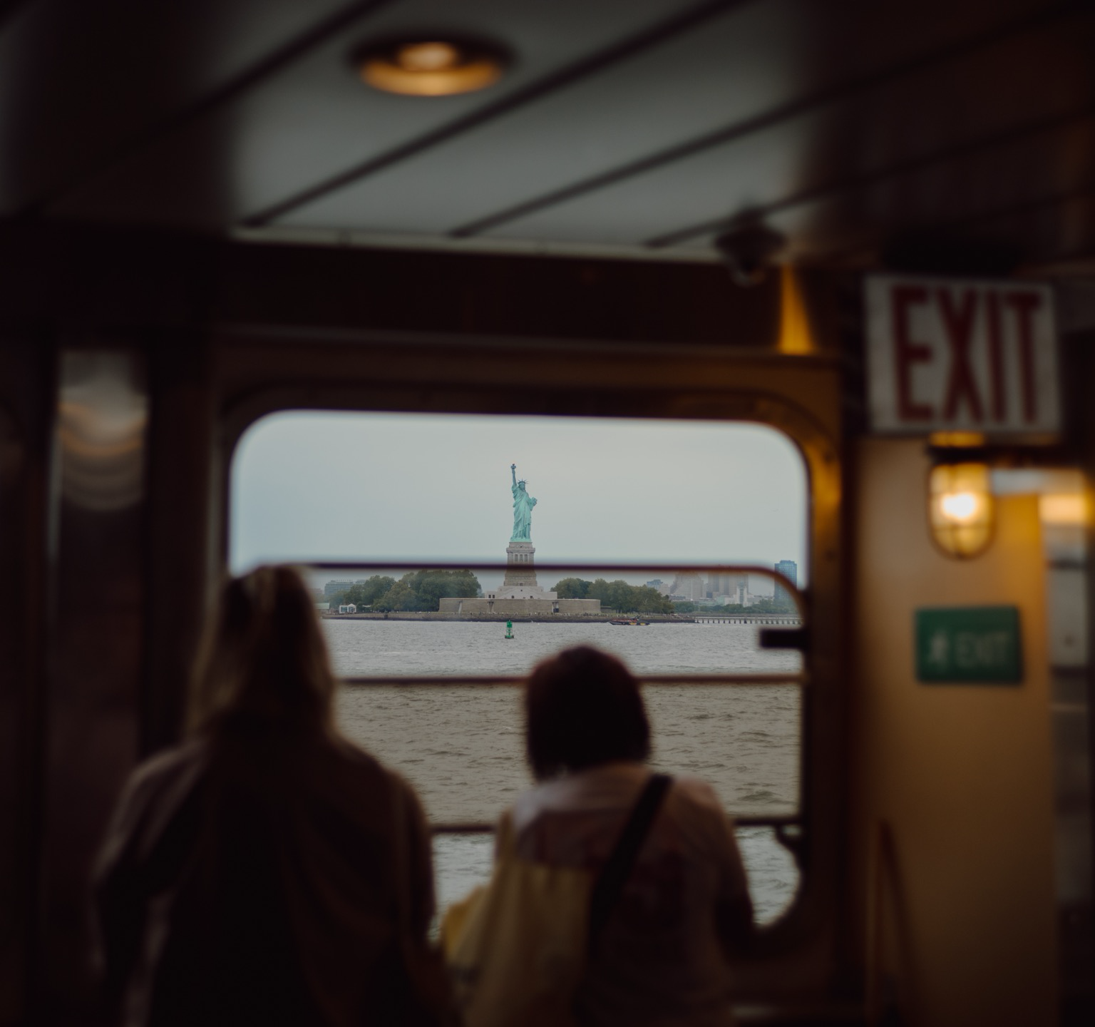 Two people seen from behind look through a ferry window at the Statue of Liberty across water under an overcast sky. An illuminated exit sign is visible on the right.