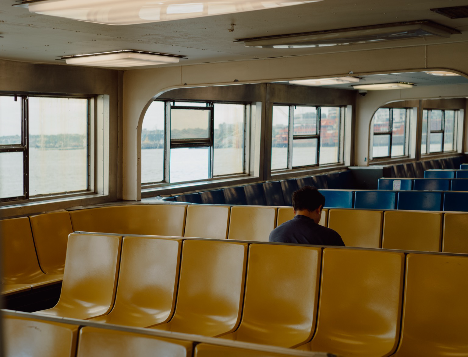 A person sits facing away in a ferry cabin with rows of yellow and blue benches. Large windows show water and distant structures under a bright sky.