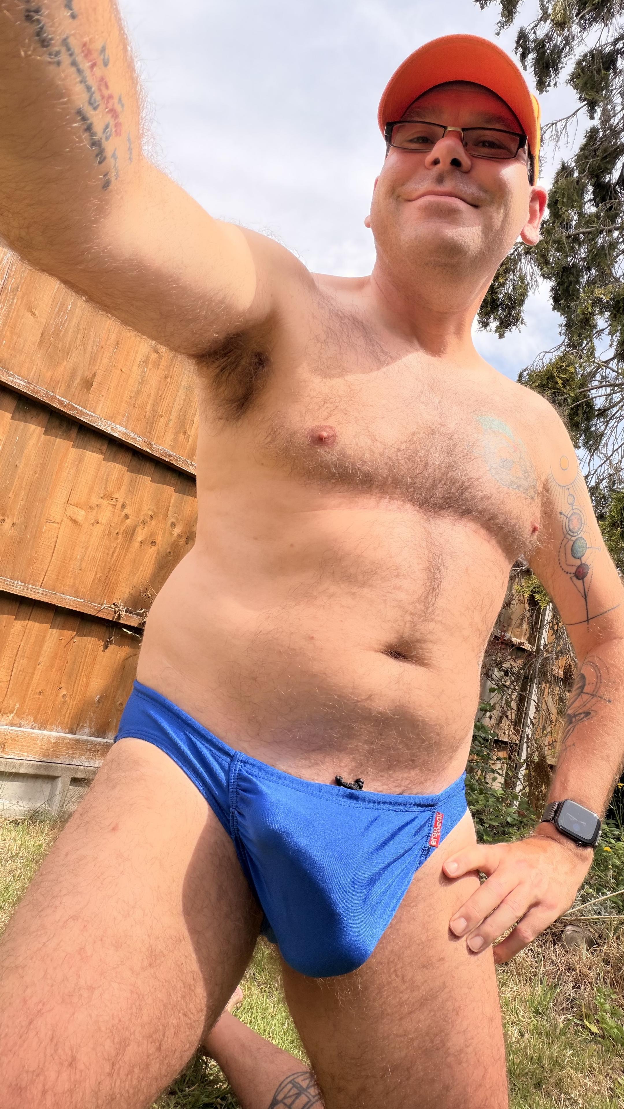 Hairy gay guy wearing royal blue ergowear swim briefs, the outline of his thick erect uncut cock point up to his right