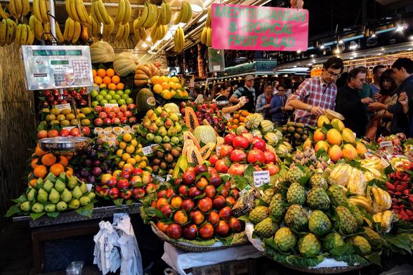 Colourful displays of fruit and vegetables at La Boqueria Market in Barcelona, including artichokes, pears, oranges, mangos, apples, and bananas.