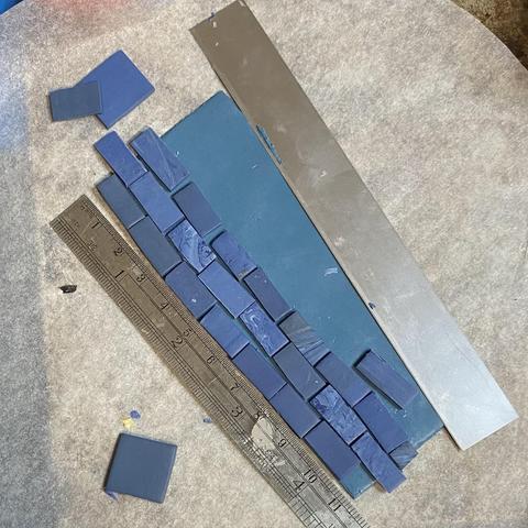 Three rows of blue tiles on a blue background sitting on a piece of baking paper. There is a miniature ruler on the left showing the whole hearth is 11cm long, and each tile 12mm long but they aren’t entirely regular as yet. A Fimo blade  lies on the other side.