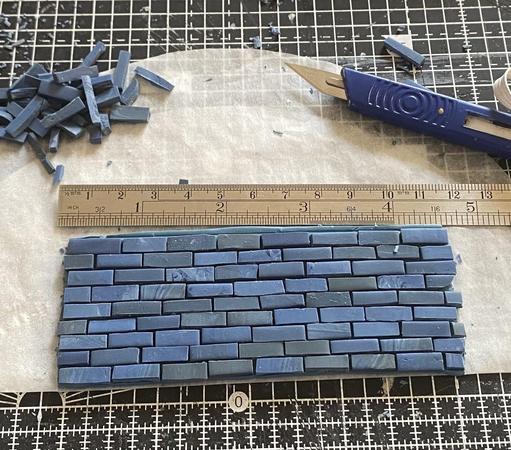 A tiled hearth for a dolls house fireplace in colours of blue A ruler lies beside showing it 11cm or just over 5 inches long. To the left is a spoil heap of broken tiles and on the right my blue craft knife.