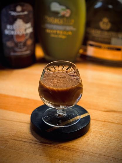 Opaque dark brown cocktail with dusted cinnamon and chocolate powder garnish in a small crystal brandy snifter sitting on a black coaster with gold band with bottles of chocolate liqueur, Botega pistachio liqueur, and cognac in the background