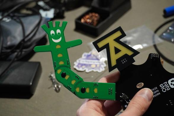 Electronic badge from the Hackaday Berline 2025 conference with two Simple Addons (SAOs) attached. The left addon is Bendy SAO.