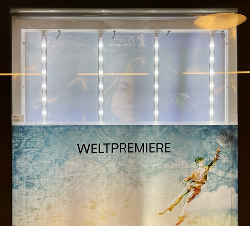 Unten ist ein Werbeplakat mit der Aufschrift Weltpremiere zu sehen. Darüber ist spiegelverkehrt ein schwach durchscheinendes Gesicht zu sehen. Davor vier Lampenketten. Hinzu kommen noch Reflexionen.