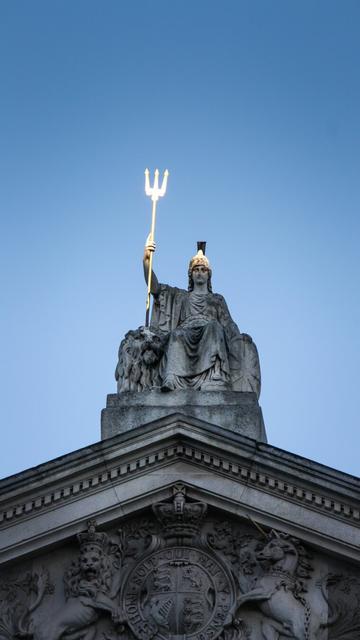 A photo of a statue of Britannia atop Somerset House. It shows a woman sitting on a throne wearing a helmet and holding up a trident. The trident is gilded, the rest of the statue is plain stone. The sun hits the statue in a way that makes the trident shine while the rest of the statue is relatively dark.