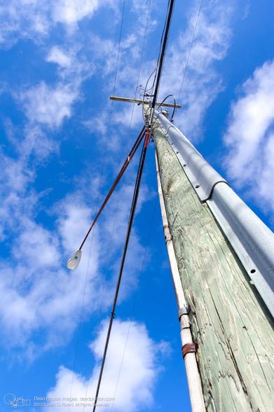 A low-angle photograph of a utility pole against a bright blue sky with scattered clouds. The pole is made of weathered wood, with a metal guard attached to one side. Power lines and a street light extend from the top of the pole, as well as communication cables.