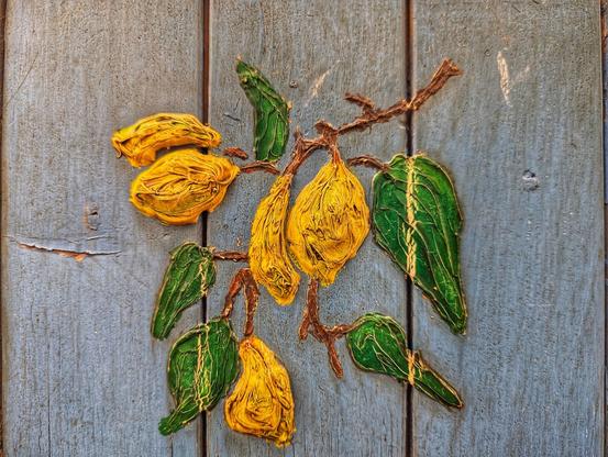 A close-up photograph shows a textured artwork of lemons and leaves attached to a weathered, light blue wooden surface. The artwork features a branch extending diagonally across the frame, bearing several bright yellow, textured lemons. Interspersed among the lemons are vibrant green leaves with textured surfaces and yellow veins. The leaves are in different orientations, some fully visible and others partially obscured by the lemons. The branch and the outlines of the lemons and leaves have a slightly raised, textured appearance, contrasting with the flatter wooden background. The wooden planks of the background are uneven in colour, showing signs of age and wear, with visible wood grain and some cracks. The lighting is even, casting soft shadows that highlight the texture of the artwork. The overall impression is of a rustic, handcrafted piece with a focus on the natural forms of lemons and foliage.