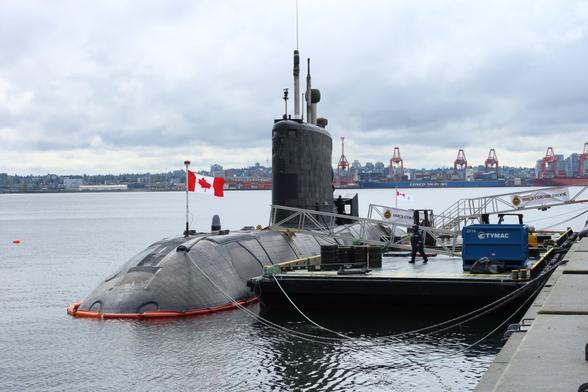 Photo of a Canadian Navy Victoria Class submarine in North Vancouver. The submarine is tied up to a barge, which is docked next to a pier. The submarine is flying a Canadian flag on the front.