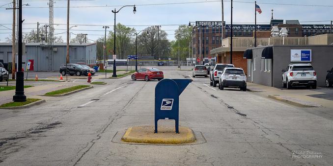 A USPS mailbox sits on a raised curb in the middle of a city street outside a post office.