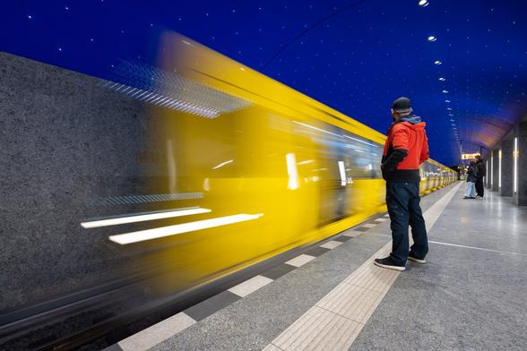 Slow motion shot of a yellow metro train entering the platform at Museumsinsel U-Bahn Station in Berlin, under a painted dark blue starry ceiling. A passenger in a bright red jacket is waiting in the foreground on the platfom adding some more color contrast to the bright yellow and dark blue tones.