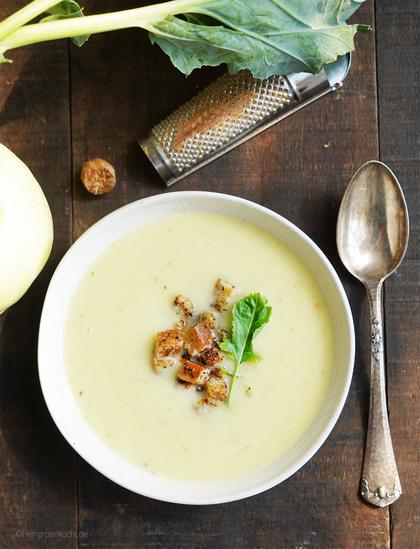 Dunkelbrauner Holzuntergrund. Darauf ein weißer, runder Suppenteller. Darin eine Kohlrabicremesuppe mit Estragoncroûtons.