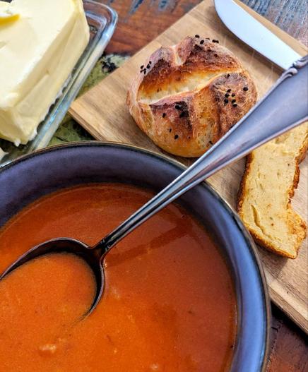 Esstisch. Graue Schale mit Tomatensuppe und silbernem Esslöffel darin. Daneben eine Butterdose mit Butter, ein Holzbrettchen mit einem schwarzkümmelbestreuten Brötchen und einem Stück Toad in the Hole mit Buttermesser.