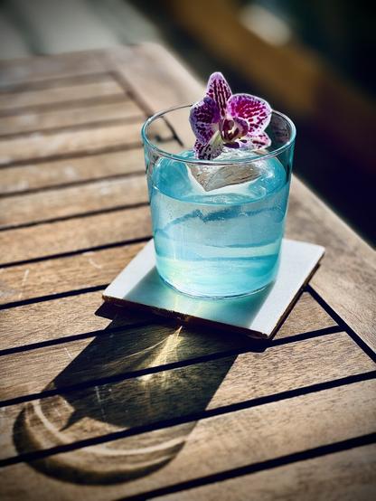 a crystal clear light blue cocktail with large rock of clear ice and white and pink orchid garnish sitting on a white coaster on a wooden table with sun shining through the drink making caustic patterns on the table