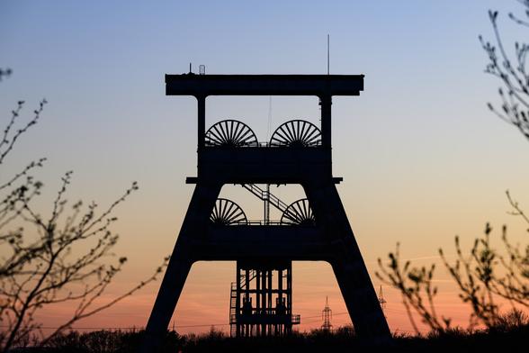 Silhouette of an old industrial mining headframe with large winding wheels, framed by tree branches at sunset; the sky transitions from a soft orange near the horizon to a deep blue at the top, creating a dramatic contrast with the dark structure.