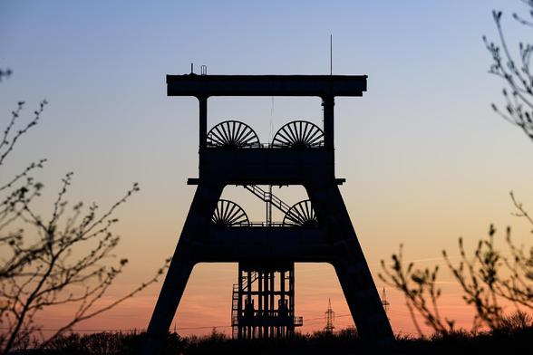 Silhouette of an old industrial mining headframe with large winding wheels, framed by tree branches at sunset; the sky transitions from a soft orange near the horizon to a deep blue at the top, creating a dramatic contrast with the dark structure.