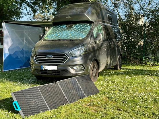 Nugget Plus camper van with extended awning, to which a sun sail is attached; a solar blanket is deployed on grass of campsite pitch