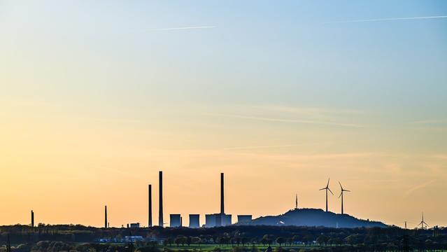Panoramic view of an industrial and renewable energy landscape at sunset, featuring silhouettes of tall smokestacks, cooling towers, and wind turbines. The sky fades from soft orange to pale blue, while the darkened horizon includes hills, transmission towers, and a scattering of buildings and trees.