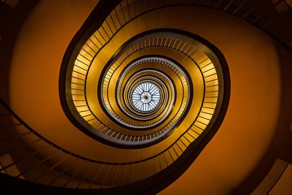 Look up view of spiral stairs in Paris with orange walls and golden electric lighting, with a  round skylight in the center at the top middle showing the blue sky and looking like an eye.
