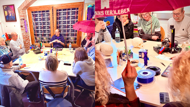 Das Bild ist eine Collage aus zwei Fotos. 

Linkes Foto: Das Bild zeigt einen Gruppenbesuch im Metalab vom Gehörlosen Sportclub Linz. Der Raum ist freundlich eingerichtet, mit Wänden, die mit Kunstwerken, Regalen voller Bücher und alten Audiokassetten geschmückt sind. Im Hintergrund ist ein in Holz eingefasstes Fenster mit einer Art Panel-Wand zu sehen.
Um einen großen, hellen Tisch herum sitzen etwa zehn Personen. Eine Person hält einen roten Regenschirm über sich und ein paar andere. 
Im Vordergrund sind verschiedene Gegenstände auf dem Tisch sichtbar: Wasserflaschen, Stifte, Papier und andere kleinere Objekte und ein 3D-Drucker.

Rechtes Foto: Das Bild zeigt eine Gruppe von Menschen, die um einen Tisch sitzen und an einem 3D-Drucker arbeiten.

Im Detail zeigt das Bild eine Gruppe von etwa sechs Personen, die um einen hellen Holztisch sitzen. Auf dem Tisch steht ein 3D-Drucker, blau-violettes Filament, ein paar 3D-gedruckte Schädel, verschiedene Kleinigkeiten wie Stifte, ein Gläschen, eine kleine Pflanze, und ein kleines, 3D-gedrucktes Otter-Tierchen. Eine Person im Vordergrund hält den Regenschirmstab.