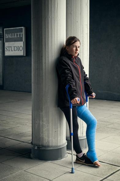 Mädchen mit blauen Gipsbein steht mit Krücken an einer Säule. Im Hintergrund ist ein Schild mit der Aufschrift „Ballett Studio“.