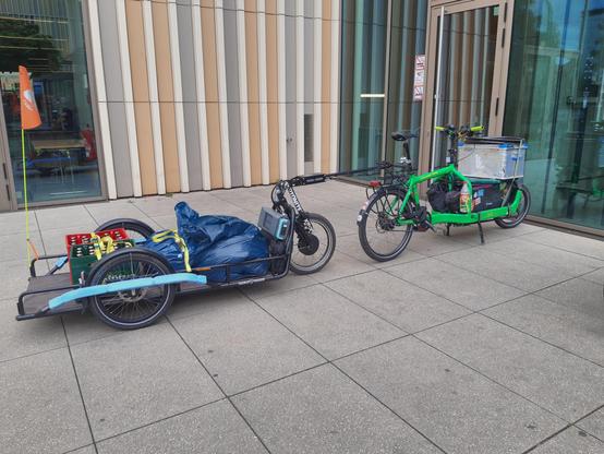 Ein Bullitt Lastenrad mit zwei Kisten vorn drauf und ein großer Carla Cargo Fahrradanhänger mit zwei Säcken und zwei Bierkisten auf der Ladefläche stehen vor einem Gebäude.