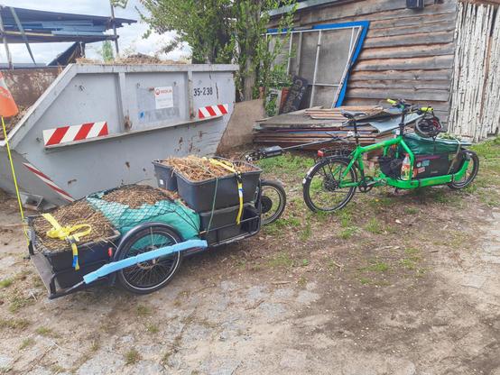Ein Bullitt Lastenrad mit einer Kiste voller Eselmist vorn drauf und ein großer Carla Cargo Fahrradanhänger mit vier Mörtelkübeln und einem großen Laubsack voller Eselmist stehen vor einen Container voller Mist
