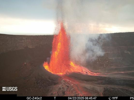 An image of the eruption in the Halema’uma’u crater this morning. The lava fountain is orange and yellow. Ribbons of red, pink, and orange flow from the base of the fountain. The rest of the scene is primarily muted browns and greys. The image is from the USGS research camera network in Hawaii Volcanoes National Park.