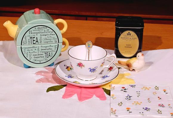 Simple tea tableau with vintage china covered in a design of tiny bouquets of roses, violets, Forget-Me-Nots, and pansies, with a matching napkin. The cup and saucer sit in the middle of a wreath of appliqued tulips on a white cloth. There is a yellow ceramic bird raising its beak in song. The small teapot is light green with yellow handles and spout and orange knob and turquoise feet; it is covered in delicate black text in various fonts using words that end with t, e, a, as puns such as affinitea, hospitalitea, generositea, creativitea, prosperitea, and so on. The tea today is Tippy Yunnan black loose leaf from Harney and Sons.