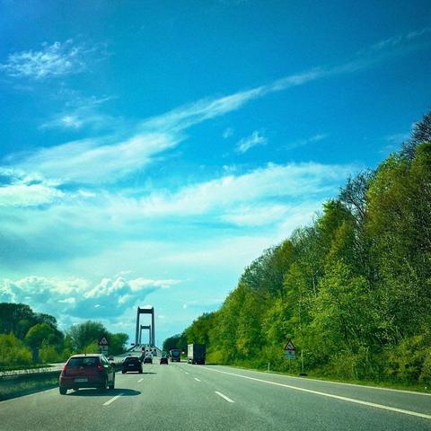 A highway scene with several cars driving toward a bridge in the distance. Lush green trees line the road, under a bright blue sky with scattered clouds.