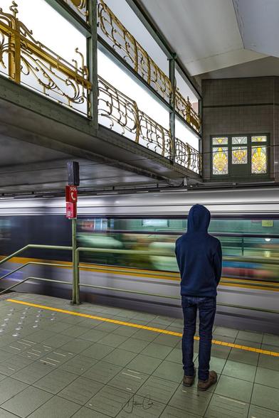 Slow exposure shot of a silhouette of a commuter seen from his back with a blurry metro train arriving on the platform in front of him in a station decorated with a golden wrought iron balustrade and stainglass windows in Art Nouveau style.
