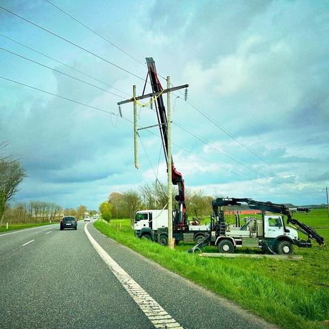 A utility vehicle and a crane are working near a power pole along a roadside. The crane is lifting a section of the pole, with nearby trees and a clear sky in the background. Cars are visible on the road.