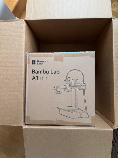 A cubical cardboard box that reads Bambu Lab A1 Mini with an outline drawing of the printer sits inside one corner of a larger shipping box