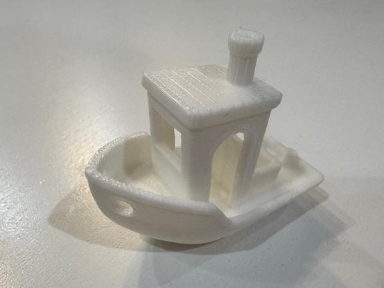 Benchy, a small white plastic 3D printed kinda cartoonish style boat sits on a white table. The bow is pointing isometrically to the left.