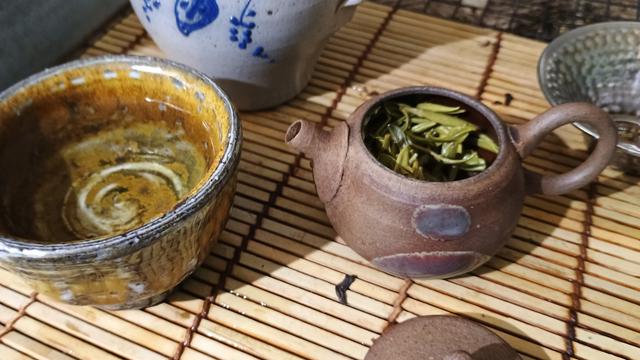 A tiny tea pot is full of green tea leaves. A little tea cup is full of green tea. There is a tiny grey stoneware pitcher in the background. Thye all sit on a bamboo sushi mat.